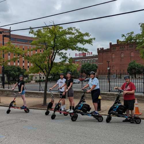 Five people on electric scooters pose together on a city street, with buildings and trees in the background.
