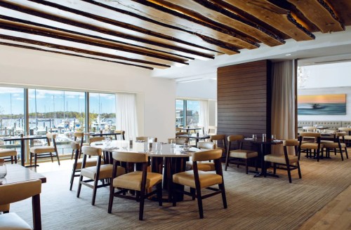 Bright restaurant interior with wooden beams, tables, and a view of a marina through large windows.