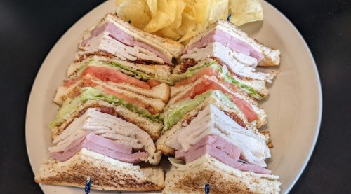 A plate of stacked sandwiches with turkey, ham, lettuce, and tomato, served with a side of potato chips.