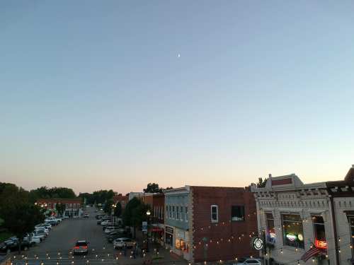 A quiet street scene at dusk, featuring shops, parked cars, and a crescent moon in a clear sky.