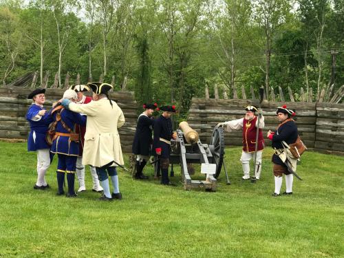 A group of people in historical military attire gather around a cannon in a grassy area with wooden fortifications.
