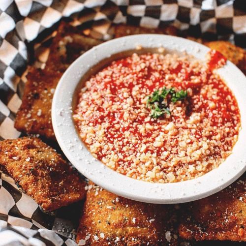 A plate of crispy fried ravioli served with marinara sauce and sprinkled with cheese, on a checkered paper background.