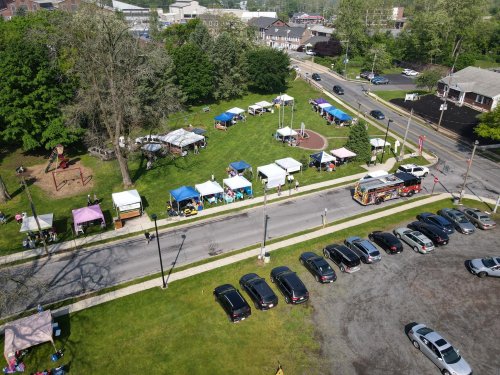 Aerial view of a park with vendor tents, a food truck, and parked cars along the street. Green trees in the background.