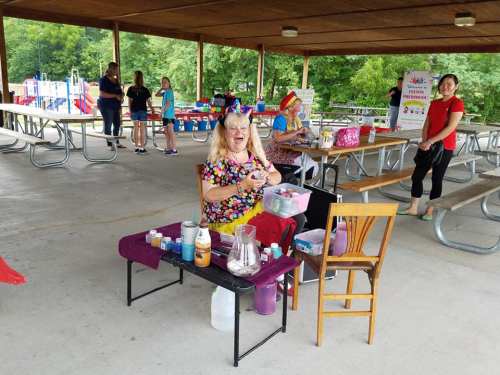 A colorful entertainer with a big smile sits at a table with craft supplies in a park pavilion, surrounded by people.