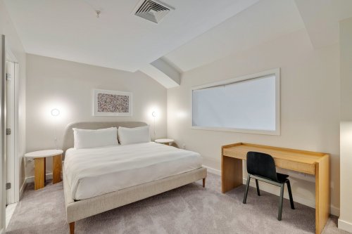 A cozy bedroom featuring a bed, two side tables, a desk, and a large window with natural light.