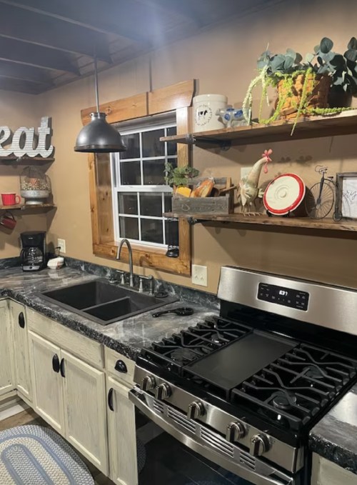 Cozy kitchen with a gas stove, double sink, wooden shelves, and rustic decor, featuring a window and plants.