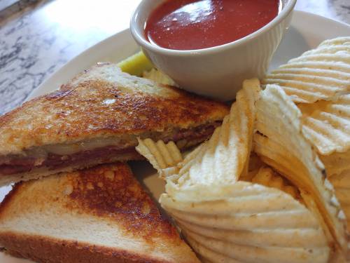 A toasted sandwich with layers of meat, served with a side of chips and a small bowl of marinara sauce.