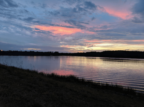 A serene lake at sunset, reflecting vibrant pink and orange hues in the sky, surrounded by trees and grassy shoreline.