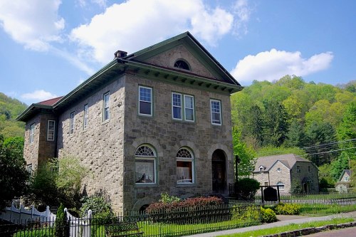 A large stone house with a green roof, surrounded by trees and a white fence, under a blue sky with fluffy clouds.