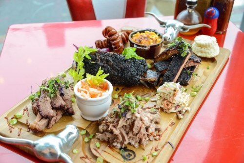 A wooden platter featuring various meats, sides, and sauces, including brisket, sausage, coleslaw, and cornbread.