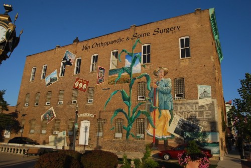 A large mural on a brick building featuring a person with corn, surrounded by framed images and text about a surgery center.