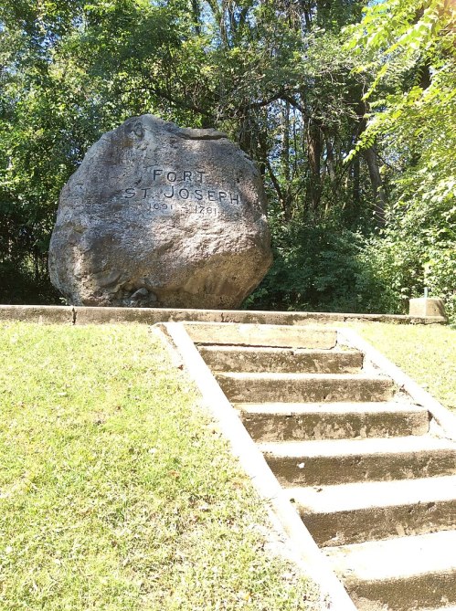 A large stone monument with "Fort St. Joseph 1686-1761" engraved, surrounded by greenery and steps leading up to it.