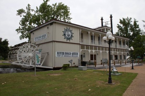 A large, two-story building resembling a riverboat, with a paddlewheel and signage for "River Road Queen" in Greenville, MS.