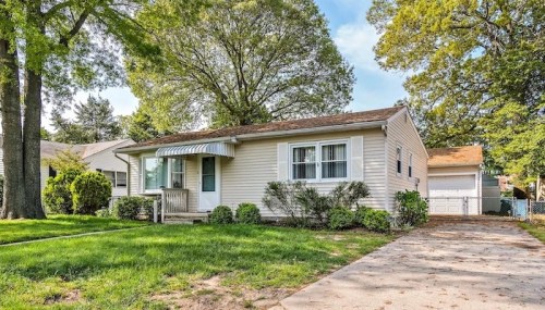 A single-story house with a front porch, surrounded by greenery and trees, on a sunny day.