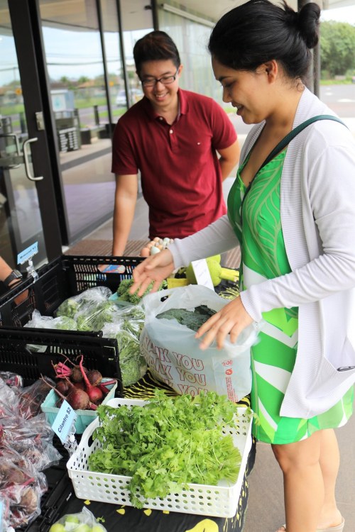 A woman in a green dress examines fresh produce at a market, while a smiling man stands nearby.
