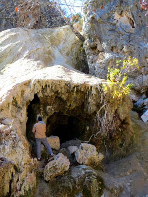 A person stands near a rocky cave entrance, surrounded by natural formations and sparse vegetation.