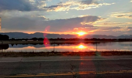 A vibrant sunset over mountains, reflecting in a calm body of water beside a road.