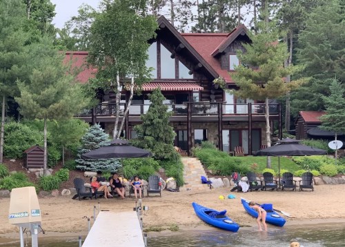 A lakeside scene featuring a large house, sandy beach, kayaks, and people relaxing under umbrellas.