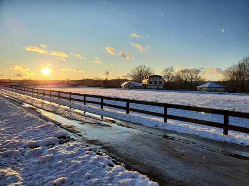 A snowy landscape at sunset, featuring a wooden fence, a road, and a few buildings in the background.