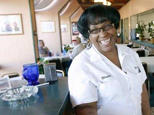 A smiling woman in a white shirt stands at a diner counter, with a cozy restaurant atmosphere in the background.