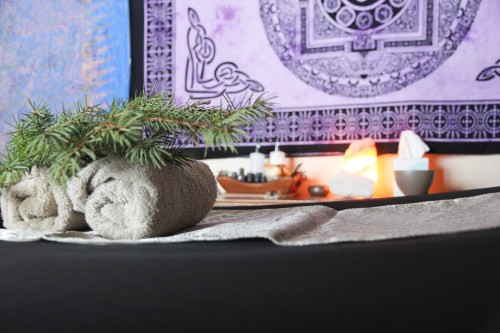 A massage table with rolled towels, a sprig of greenery, and a colorful tapestry in the background.