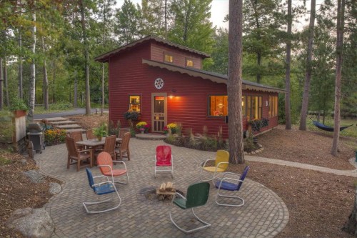 A cozy red cabin surrounded by trees, with a fire pit and colorful chairs in a stone patio area.