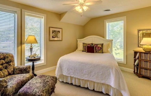 Cozy bedroom with a white bed, floral accents, armchair, and large windows letting in natural light.