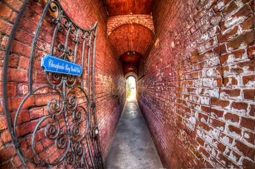 A narrow brick corridor with a decorative gate, leading to a bright light at the end.