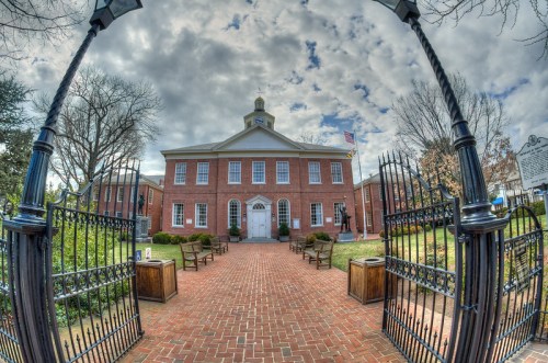 Historic building with a clock tower, surrounded by a garden and benches, under a cloudy sky.
