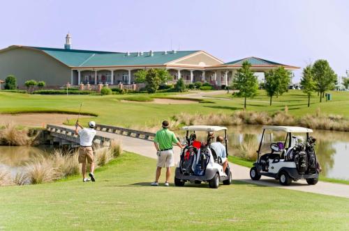 A golfer swings on a lush course, while two others watch from golf carts near a pond and clubhouse in the background.