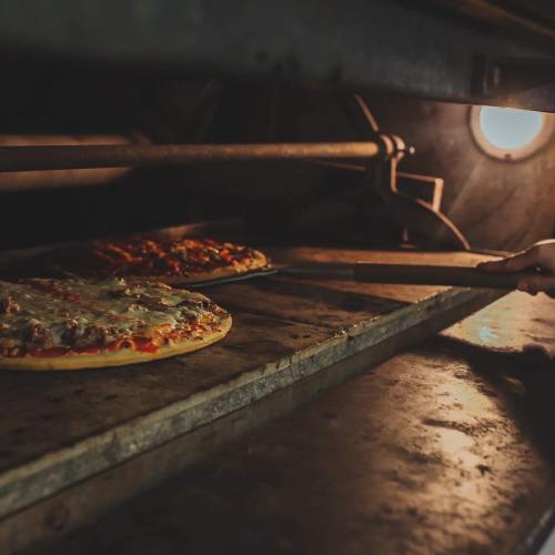 A hand pulls a pizza from a stone oven, with another pizza baking in the background. Warm, inviting lighting.