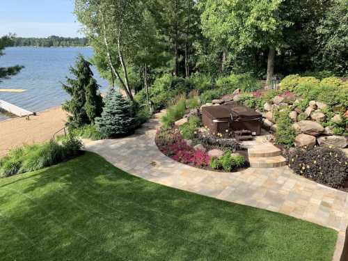 A scenic lakeside garden featuring a hot tub, stone pathway, colorful flowers, and lush greenery by the water.