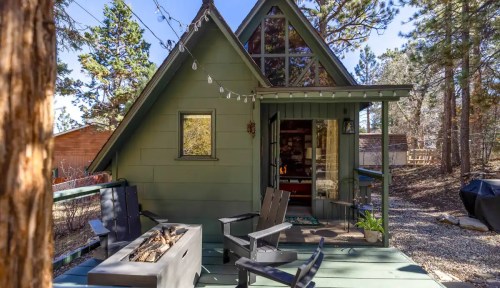 A cozy green A-frame cabin with a porch, surrounded by trees and outdoor seating, featuring string lights and a fire pit.