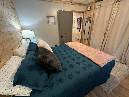 Cozy bedroom featuring a bed with teal bedding, a pink throw, wooden walls, and a lamp beside the bed.