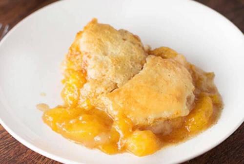 A serving of peach cobbler with golden biscuit topping on a white plate, surrounded by warm peach filling.
