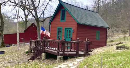 A small red cabin with green trim, a wooden deck, and an American flag, surrounded by trees and grassy land.
