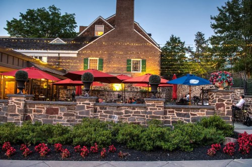 A cozy restaurant patio with red umbrellas, stone walls, and colorful flowers, illuminated by string lights at dusk.