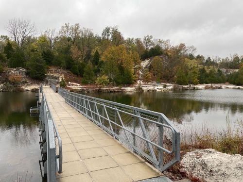 A metal bridge leads over a calm lake, surrounded by trees and rocky terrain under a cloudy sky.