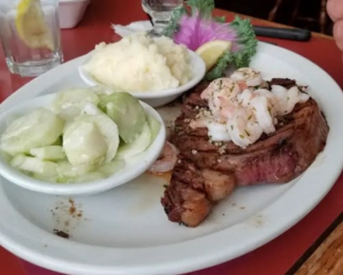 A plate featuring a grilled steak topped with shrimp, served with mashed potatoes and a side of cucumber salad.
