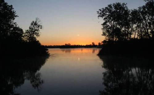 Serene sunset over a calm river, framed by silhouettes of trees on either side.