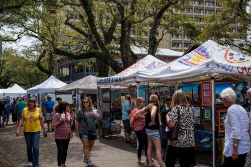 A bustling outdoor art market with vendors under tents and visitors browsing artwork on a sunny day.