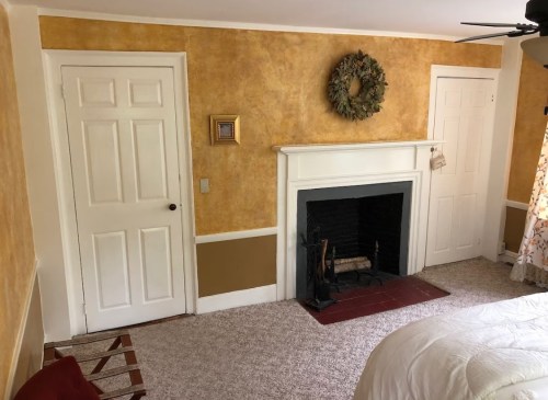 Cozy room with yellow walls, a fireplace, and two doors. A wreath hangs above the mantel, and a patterned curtain is visible.