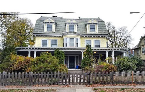 A large, historic yellow house with a steep roof, surrounded by greenery and a wooden fence.