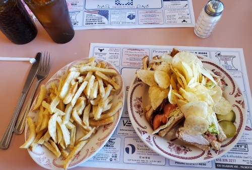 A plate with a sandwich topped with chips, pickles, and a side of golden French fries on a pink table.