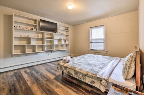 A cozy bedroom featuring a bed, shelves with decor, a TV, and wooden flooring, illuminated by natural light.