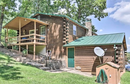 A rustic log cabin with a green metal roof, porch, and satellite dish, surrounded by trees and grass.