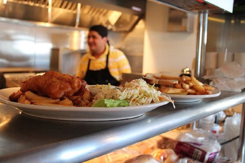 A plate of fried fish and chips with coleslaw, served in a restaurant kitchen with a chef in the background.