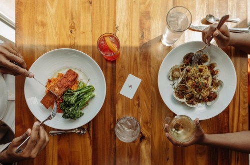 Two plates of food on a wooden table, one with salmon and broccoli, the other with pasta and clams, alongside drinks.