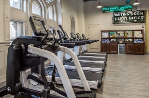 A row of treadmills in a modern gym, with a ticket office and waiting room sign in the background.