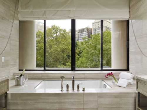 A modern bathroom with a large tub, towels, and a view of greenery through large windows.